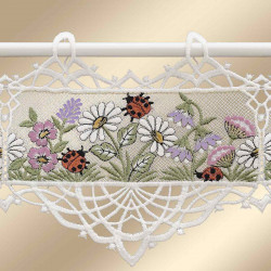 An einer Stange hängt ein gesticktes Paneel mit weißen Gänseblümchen, lila Blüten und grünen Blättern. Mehrere rote Marienkäfer mit schwarzen Punkten sind zwischen den floralen Elementen platziert. Die Ränder des Paneels sind mit detaillierter weißer Spitze abgeschlossen.
