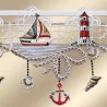 Die gestickte Spitzenborte zeigt ein Segelboot und einen rot gestreiften Leuchtturm über welligem Wasser vor einem netzgemusterten Hintergrund. Zu den hängenden Elementen gehören Muscheln, ein roter Anker und eine Muschelschale, die alle auf einem beigen Hintergrund präsentiert werden.