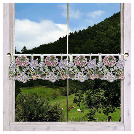 Ein Fenster mit einem durchsichtigen Spitzenvorhang mit Blumenmustern in Rosa, Violett und Gelb, das den Blick auf grüne Hügel, Bäume und grasende Kühe freigibt.