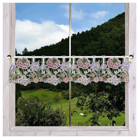Ein Fenster mit einem durchsichtigen Spitzenvorhang mit Blumenmustern in Rosa, Violett und Gelb, das den Blick auf grüne Hügel, Bäume und grasende Kühe freigibt.