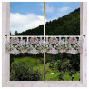 Ein Fenster mit einem durchsichtigen Spitzenvorhang mit Blumenmustern in Rosa, Violett und Gelb, das den Blick auf grüne Hügel, Bäume und grasende Kühe freigibt.
