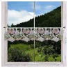 Ein Fenster mit einem durchsichtigen Spitzenvorhang mit Blumenmustern in Rosa, Violett und Gelb, das den Blick auf grüne Hügel, Bäume und grasende Kühe freigibt.