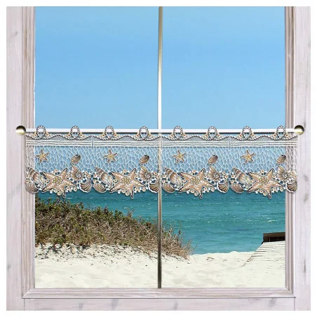 Ein Spitzenvorhang mit einem Muschel- und Seesternmuster hängt an einer Stange über einem Fenster. Außerhalb des Fensters sind Sanddünen, Gras und ein blauer Ozean mit sanften Wellen unter einem klaren Himmel zu sehen. Der Fensterrahmen ist aus hellem Holz gefertigt.