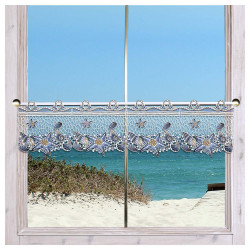 Ein Fenster mit weißer Umrandung hat einen Spitzenvorhang, der mit Seestern- und Muschelmustern verziert ist. Durch das Fenster sind Sanddünen, grüne Sträucher und das blaue Meer unter einem klaren Himmel zu sehen, was eine Küstenlandschaft darstellt.