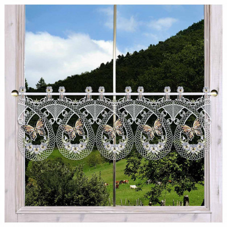 Ein Fenster mit einem hellen Holzrahmen wird von einem weißen Spitzenvorhang verdeckt, der mit detaillierten Schmetterlings- und Blumenmotiven verziert ist. Durch das Fenster sieht man einen grünen Hügel mit Bäumen, grasenden Kühen und einem blauen Himmel mit vereinzelten weißen Wolken.