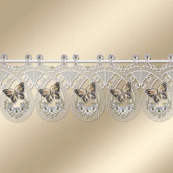 Spitzenvolant mit sich wiederholenden ovalen Motiven, die jeweils einen Schmetterling und kleine weiße Blüten mit grünen Blättern enthalten. Die Spitze ist aufwändig genäht und mit Wellenkanten versehen. Er wird an einer Stange hängend vor einem beigen Hintergrund präsentiert.