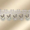 Spitzenvolant mit sich wiederholenden ovalen Motiven, die jeweils einen Schmetterling und kleine weiße Blüten mit grünen Blättern enthalten. Die Spitze ist aufwändig genäht und mit Wellenkanten versehen. Er wird an einer Stange hängend vor einem beigen Hintergrund präsentiert.