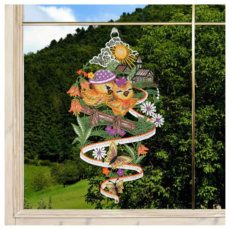 Buntes Spitzenornament hängt in einem Fenster, auf dem zwei Vögel auf einem Zaun, Blumen, Schmetterlinge, eine Sonne und ein kleines Haus zu sehen sind. Grüne Hügel, Bäume und blauer Himmel sind durch das Fenster im Hintergrund zu sehen.