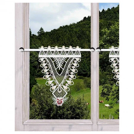 Zarte Spitzenvorhänge mit aufwendigen Mustern hängen an einem Fenster mit Holzrahmen, durch das der Blick auf eine üppige, grüne Landschaft mit Bäumen und grasenden Kühen fällt. Eine kleine, bunte Blume ziert die Spitze der Gardine.