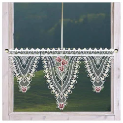 Ein Fenster mit beigem Rahmen ist mit einem kunstvollen Spitzenvolant geschmückt. Die Spitze weist dreieckige Muster mit roten Blumenmotiven und zarten weißen Details auf und gibt den Blick auf eine verschwommene grüne Landschaft draußen frei.