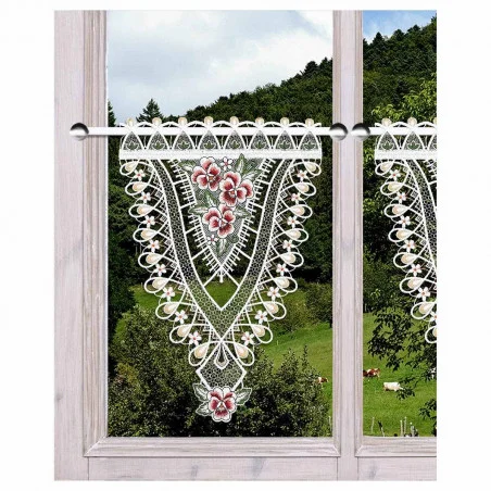 Ein Holzfenster zeigt einen Spitzenvolant mit floralen Mustern. Der Blick nach draußen zeigt eine üppig grüne Landschaft mit Bäumen und einigen grasenden Tieren auf einem Hügel.