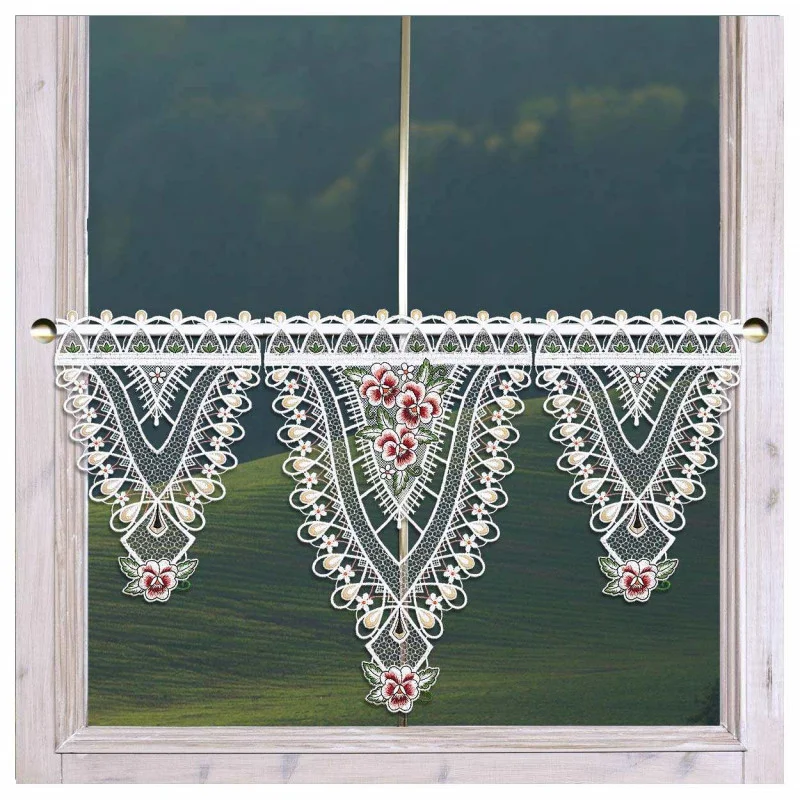 Ein Fenster mit beigem Rahmen ist mit einem kunstvollen Spitzenvolant geschmückt. Die Spitze weist dreieckige Muster mit roten Blumenmotiven und zarten weißen Details auf und gibt den Blick auf eine verschwommene grüne Landschaft draußen frei.