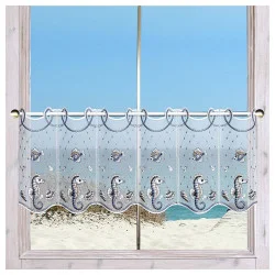 Ein Fenster mit einem durchsichtigen, dekorativen Vorhang mit Fisch- und Seepferdchenmotiven mit Blick auf einen Sandstrand und den ruhigen blauen Ozean unter einem klaren Himmel.