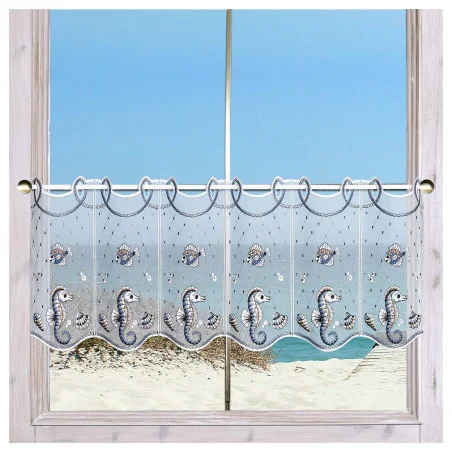Ein Fenster mit einem durchsichtigen, dekorativen Vorhang mit Fisch- und Seepferdchenmotiven mit Blick auf einen Sandstrand und den ruhigen blauen Ozean unter einem klaren Himmel.