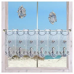 Ein Fenster mit transparenten, dekorativen Vorhängen mit Seepferdchen- und Fischmotiven bietet Blick auf einen Sandstrand und das blaue Meer. Zwei gestickte Fensterbilder mit Meeresmotiven hängen am Fenster.