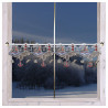 Weiß gerahmtes Fenster mit einem dekorativen maritimen Gardine-Spitzenvolant mit Ankern, Vögeln und Muscheln. Draußen entfaltet sich eine verschneite, bewaldete Winterlandschaft unter einem dämmrig-blauen Himmel.