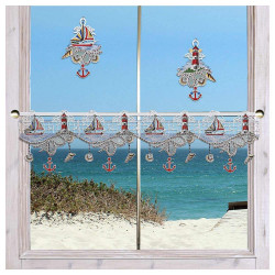 Ein Fenster ist mit der Scheibengardine „Meeresbrise“ im maritimen Stil geschmückt. Sie zeigt rote Anker, Leuchttürme, Boote und Muscheln aus zarter Vogtlädischer Spitze. Draußen erstrecken sich Sanddünen und blaue Meereswellen unter einem klaren Himmel.