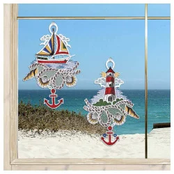 Zwei Fensterbilder mit nautischem Thema – ein Segelboot und ein Leuchtturm mit Ankern – bringen einen Hauch von Bad-Deko und Meeresbrise an Ihr Fenster mit Blick auf einen Sandstrand, grüne Sträucher und das glitzernde Meer unter einem blauen Himmel.