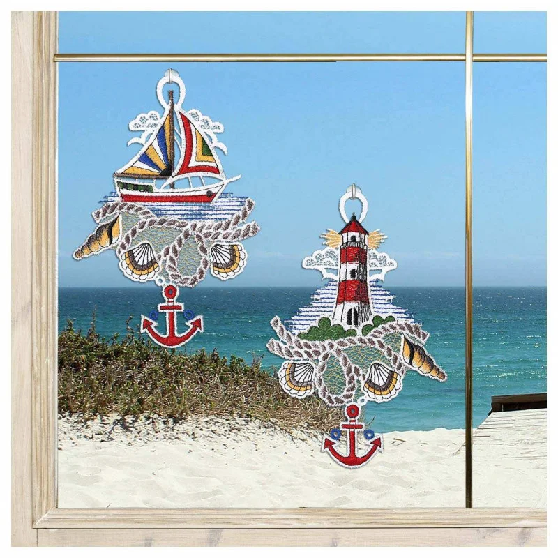 Zwei Fensterbilder mit nautischem Thema – ein Segelboot und ein Leuchtturm mit Ankern – bringen einen Hauch von Bad-Deko und Meeresbrise an Ihr Fenster mit Blick auf einen Sandstrand, grüne Sträucher und das glitzernde Meer unter einem blauen Himmel.