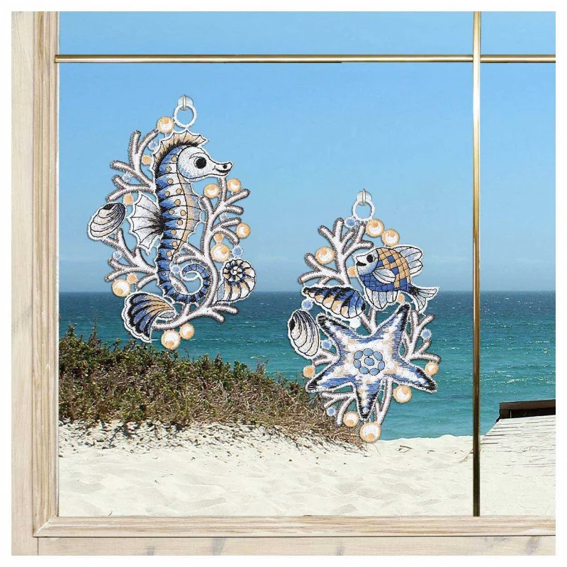 Zwei gestickte maritime Fensterbilder, eines in Form eines Seepferdchens und das andere in Form eines Fisches, hängen an einem Fenster mit Blick auf einen Sandstrand und das Meer unter einem klaren blauen Himmel.