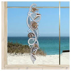 Ein dekoratives Spitzen-Fensterbild mit vogtländischer Spitze mit Fischen, Muscheln und Meeresmotiven hängt in einem Fenster mit Blick auf einen Sandstrand, Meereswellen und einen klaren blauen Himmel.