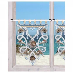 An einem Fenster hängt ein dekorativer Spitzenvorhang der Vogtländischen Spitze mit einem maritimen Muster aus Fischen, Muscheln und Meereswellen. Draußen sieht man durch den maritimen Scheibenhänger einen Sandstrand, blaues Meer und klaren Himmel.
