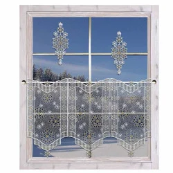 Ein Fenster mit frostigen Schneeflocken-Fensterbildern und Spitzenvorhängen mit Schneeflockenmuster, das eine verschneite Landschaft mit Bäumen und einem klaren blauen Himmel zeigt.