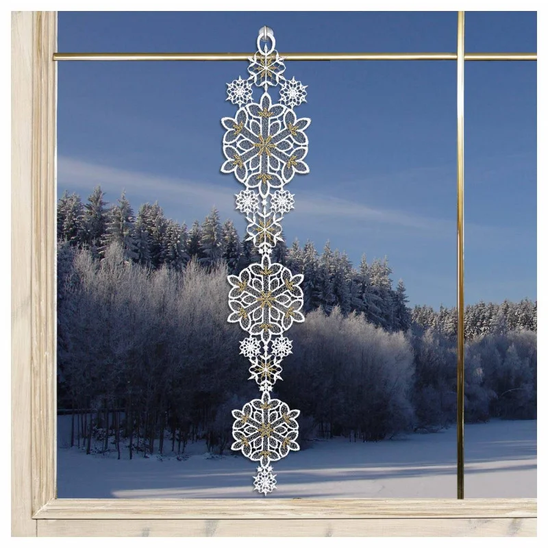 Eine dekorative Giralnde aus gestickten weißen Schneeflocken hängt in einem Fenster, aus dem ein verschneiter Wald und ein blauer Himmel zu sehen sind.