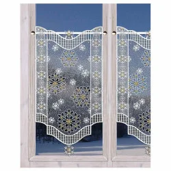 Ein weißer Spitzenvorhang mit goldenen und weißen Schneeflockenmustern hängt an einer Stange über einem Fenster. Der Fensterrahmen ist aus hellem Holz, und im Hintergrund ist eine verschneite Szene im Freien zu sehen.