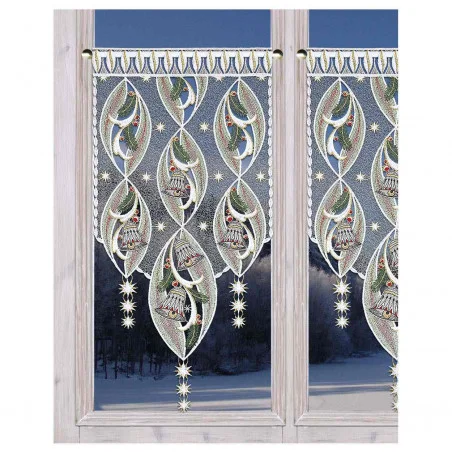 Dekorative Spitzenvorhänge mit verschlungenen Glocken und Tannenzweigen hängen an einem Fenster und geben den Blick auf eine verschneite Winterlandschaft und dunkle Bäume frei.