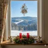 Eine festliche Fensterszene mit einem verschneiten Wald-Wintermotiv draußen, einem Fensterbild Stern am Fenster und einem Adventskranz mit vier roten Kerzen, Grünzeug und Figuren auf der Fensterbank.
