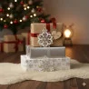 Drei eingepackte Weihnachtsgeschenke, geschmückt mit Bändern und Schneeflockenornamenten, sitzen auf einem flauschigen Teppich. Im gemütlichen Hintergrund verstärken ein beleuchteter Weihnachtsbaum und eine leuchtende Lampe das Winterflair der Szene.