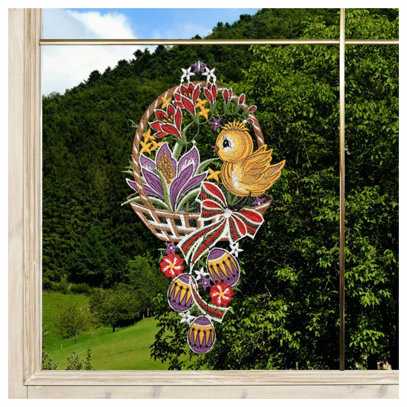 Eine farbenfrohe gestickte Fensterdekoration mit einem gelben Küken in einem geflochtenen Korb mit roten Blumen, lila Tulpen, grünen Blättern und hängenden roten Ornamenten mit Schleifen. Durch ein Fenster gesehen vor einer bewaldeten Berglandschaft.