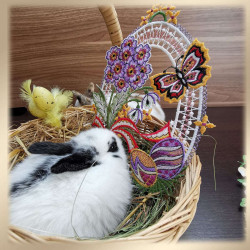 Ein schwarz-weißer Hase ruht in einem mit Stroh ausgekleideten Weidenkorb. Der Korb enthält bunte Ostereier, verschiedene Blumen, ein dekoratives gelbes Küken und eine hellviolette Fensterbild Osterei mit Schmetterlingsmotiv. Der Hintergrund besteht aus Holzplatten.