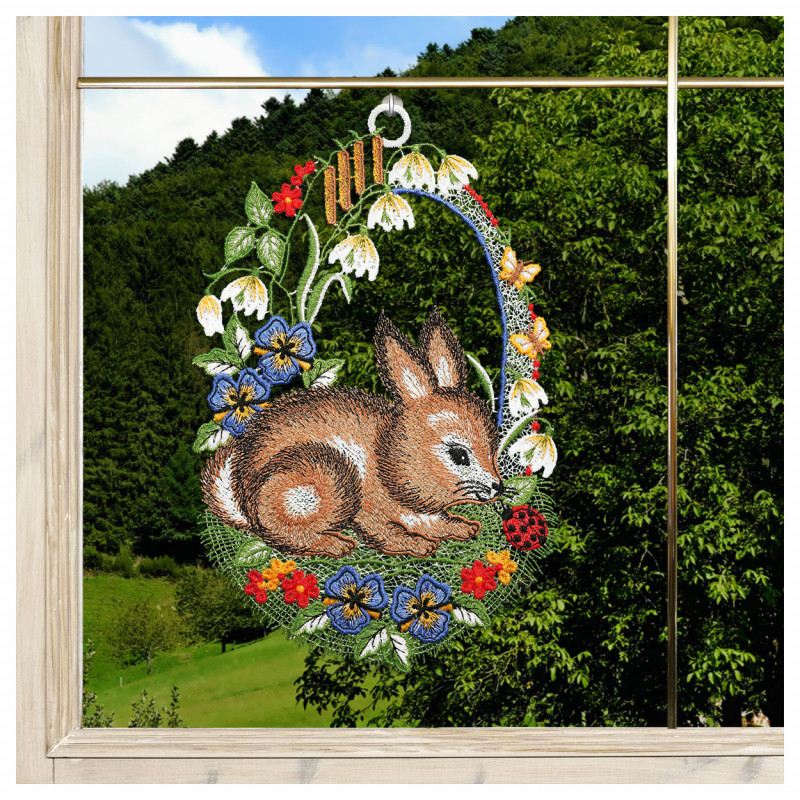 Das Fensterbild Hase mit Käfer zeigt einen gestickten braunen Hasen inmitten von blauen, gelben und weißen Blumen. Ein Marienkäfer befindet sich in der Nähe der Pfote des Kaninchens, das vor grünen Hügeln und einem teilweise bewölkten blauen Himmel zu sehen ist.