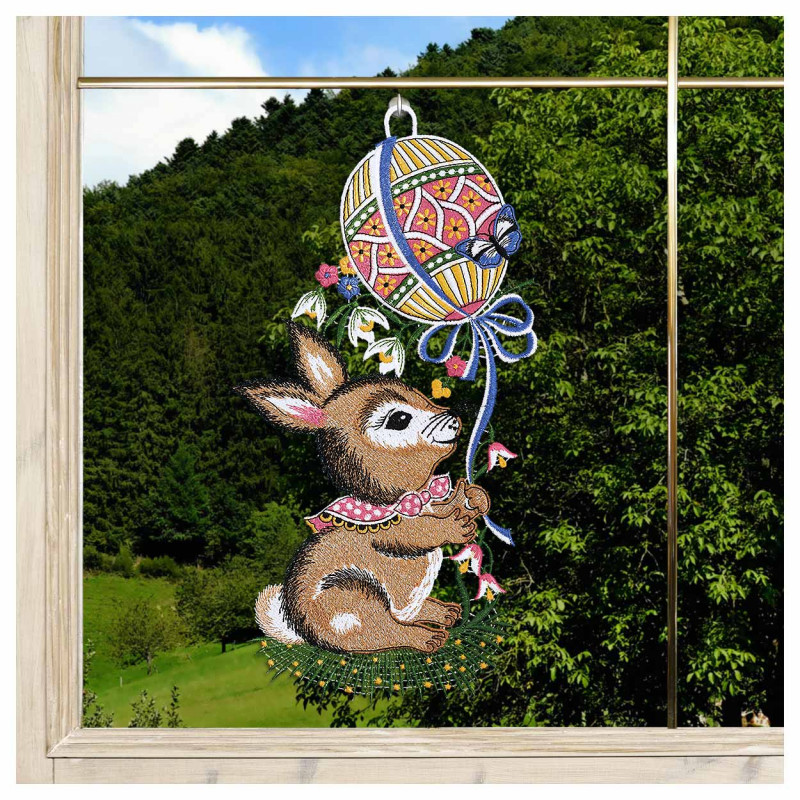 Das "Fensterbild Hase mit Osterballon vogtländischer Spitze" (34x18 cm) ist eine Fensterdekoration, die einen braunen Hasen mit rosa Schal darstellt, der im Gras sitzt und einen Osterballon in der Hand hält, gefertigt im Stil vogtländischer Spitze.
