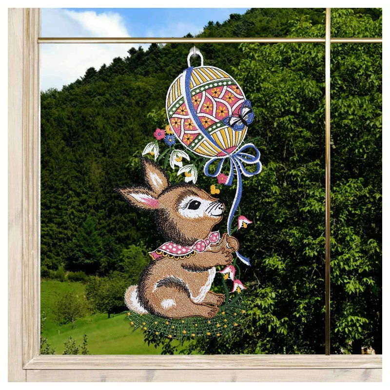 Das "Fensterbild Hase mit Osterballon vogtländischer Spitze" (34x18 cm) ist eine Fensterdekoration, die einen braunen Hasen mit rosa Schal darstellt, der im Gras sitzt und einen Osterballon in der Hand hält, gefertigt im Stil vogtländischer Spitze.