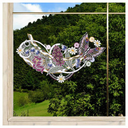 Das Fensterbild Frühlingssinfonie Plauener Spitze mit Blumengirlande ist auf einer Fensterscheibe abgebildet, die ein buntes Blumen- und Schmetterlingsmuster mit lila, rosa und weißen Blüten und grünen Blättern zeigt. Draußen stehen grüne Bäume und ein grasbewachsener Hügel.