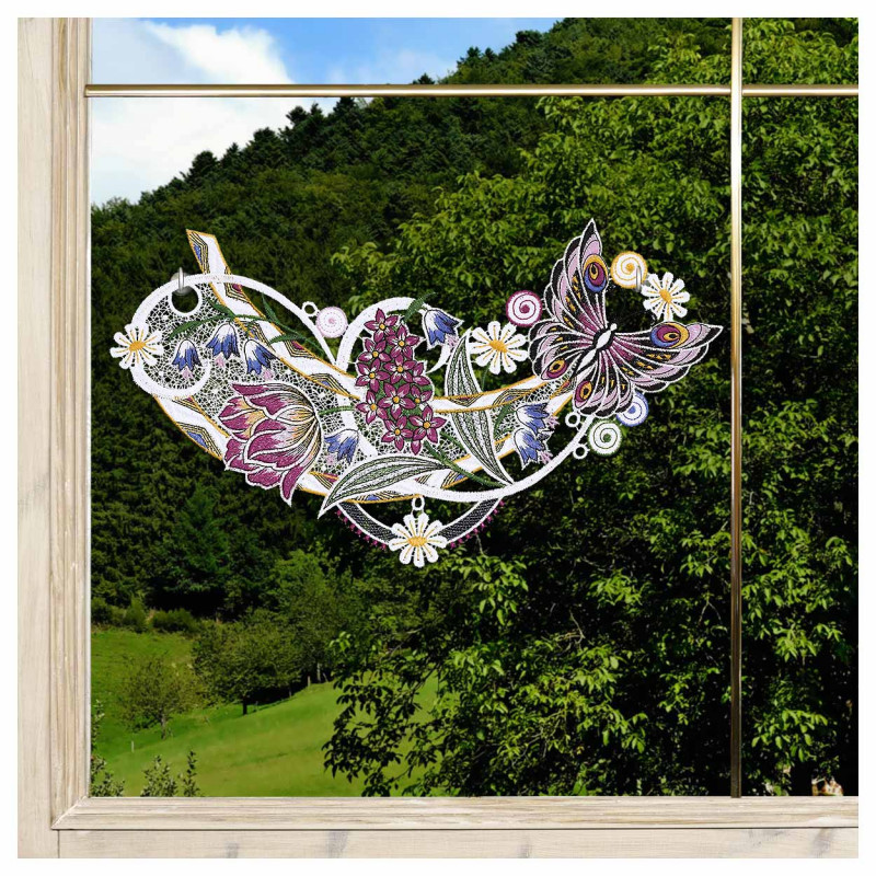 Das Fensterbild Frühlingssinfonie Plauener Spitze mit Blumengirlande ist auf einer Fensterscheibe abgebildet, die ein buntes Blumen- und Schmetterlingsmuster mit lila, rosa und weißen Blüten und grünen Blättern zeigt. Draußen stehen grüne Bäume und ein grasbewachsener Hügel.