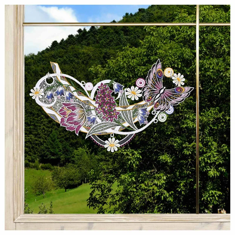 Das Fensterbild Frühlingssinfonie Plauener Spitze mit Blumengirlande ist auf einer Fensterscheibe abgebildet, die ein buntes Blumen- und Schmetterlingsmuster mit lila, rosa und weißen Blüten und grünen Blättern zeigt. Draußen stehen grüne Bäume und ein grasbewachsener Hügel.