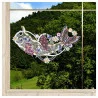 Das Fensterbild Frühlingssinfonie Plauener Spitze mit Blumengirlande ist auf einer Fensterscheibe abgebildet, die ein buntes Blumen- und Schmetterlingsmuster mit lila, rosa und weißen Blüten und grünen Blättern zeigt. Draußen stehen grüne Bäume und ein grasbewachsener Hügel.