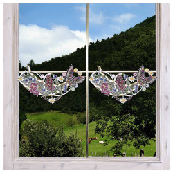 Ein Fenster mit durchsichtigen Spitzenvorhängen, Plauener Spitze "Frühlingssinfonie", zeigt eine Blumengirlande und zwei gestickte Vögel, die sich gegenüberstehen. Draußen ist ein grüner Hügel mit Bäumen, Gras und Kühen unter einem teilweise bewölkten Himmel zu sehen.