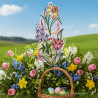 Ein Fensterbild Frühlingsfreude Plauener Spitze mit floralen Motiven in Form einer großen verschnörkelten "8" steht aufrecht im grünen Gras. In der Nähe steht ein Weidenkorb mit pastellfarbenen Ostereiern, Tulpen, Narzissen und Glockenblumen auf einer sonnenbeschienenen Frühlingswiese.