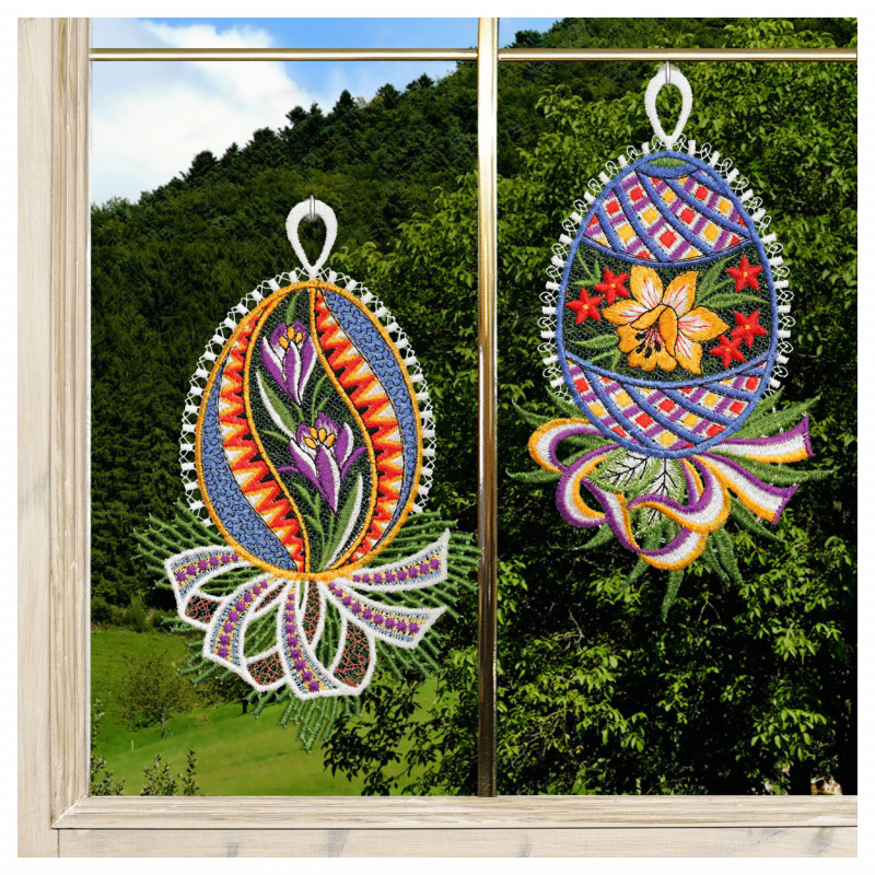 Zwei gestickte, eiförmige Ornamente aus dem Fensterbilder 2er Set Ostereier mit Krokus und Narzisse Plauener Spitze blau hängen in einem Fenster. Jedes Ornament ist mit blauen Krokus- und Narzissenblüten verziert. Draußen sind grüne Bäume und Hügel unter einem klaren Himmel zu sehen.