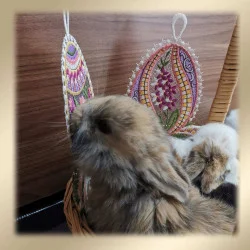 Ein flauschiges braunes Kaninchen mit langen Ohren sitzt in einem Korb und schnüffelt an einem bunten, gestickten eiförmigen Ornament. Neben dem Korb befindet sich ein weiterer Plüschhase, und der Hintergrund besteht aus einer Holzoberfläche mit weichen, beigen Vignettenrändern.