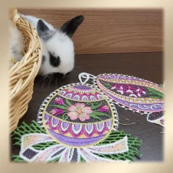 Ein schwarz-weißer Hase lugt aus einem geflochtenen Korb neben zwei aufwändig bestickten, bunten Ostereiern mit floralen und geometrischen Mustern hervor, die auf einer dunklen Holzfläche liegen. Das Bild hat einen weichen beigen Rand.