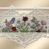 Ein Wandbehang aus Spitze mit gestickten Gänseblümchen, lila und rosa Blumen, grünen Blättern und mehreren roten und schwarzen Marienkäfern. Der Wandbehang ist mit einer detaillierten weißen Spitzenbordüre versehen und wird an einer weißen Stange mit drei Schlaufen aufgehängt.