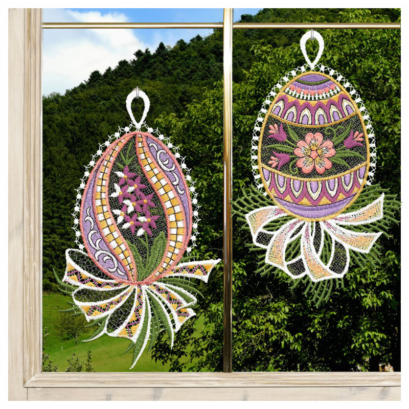 Zwei gestickte Ostereier, verziert mit floralen und geometrischen Mustern in verschiedenen Farben, hängen an einem Fenster. Jedes Ei hat eine weiße Spitzenschleife und eine bandartige Spitze am unteren Ende. Durch das Fenster sind grüne Hügel, Bäume, blauer Himmel und Wolken zu sehen.