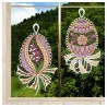Zwei gestickte Ostereier, verziert mit floralen und geometrischen Mustern in verschiedenen Farben, hängen an einem Fenster. Jedes Ei hat eine weiße Spitzenschleife und eine bandartige Spitze am unteren Ende. Durch das Fenster sind grüne Hügel, Bäume, blauer Himmel und Wolken zu sehen.