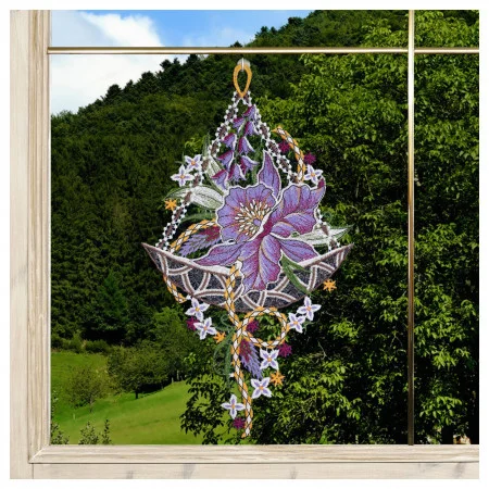 Ein Spitzenornament mit einer auffälligen violetten Blüte und verschlungenen Mustern hängt in einem Fenster. Durch das Glas ist ein dicht mit grünen Bäumen bewachsener Hügel unter einem klaren Himmel zu sehen, der einen lebendigen natürlichen Hintergrund für die Dekoration bildet.